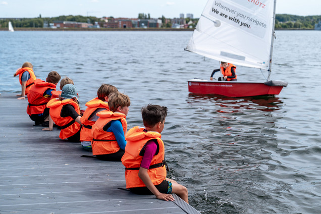 Kinder sitzen mit Kinderwesten auf dem Steg und ein Kind sitzt im Opti in einem Kurs vom Segelcamp Kiel