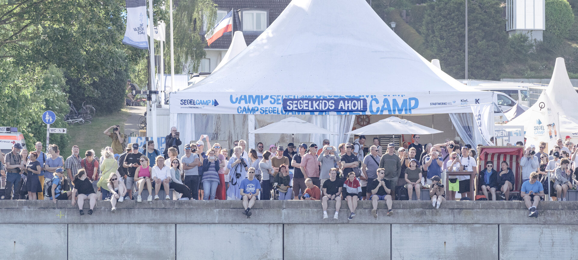 Menschenmenge steht/sitzt an der Kiellinie und beobachten ein Segelevent